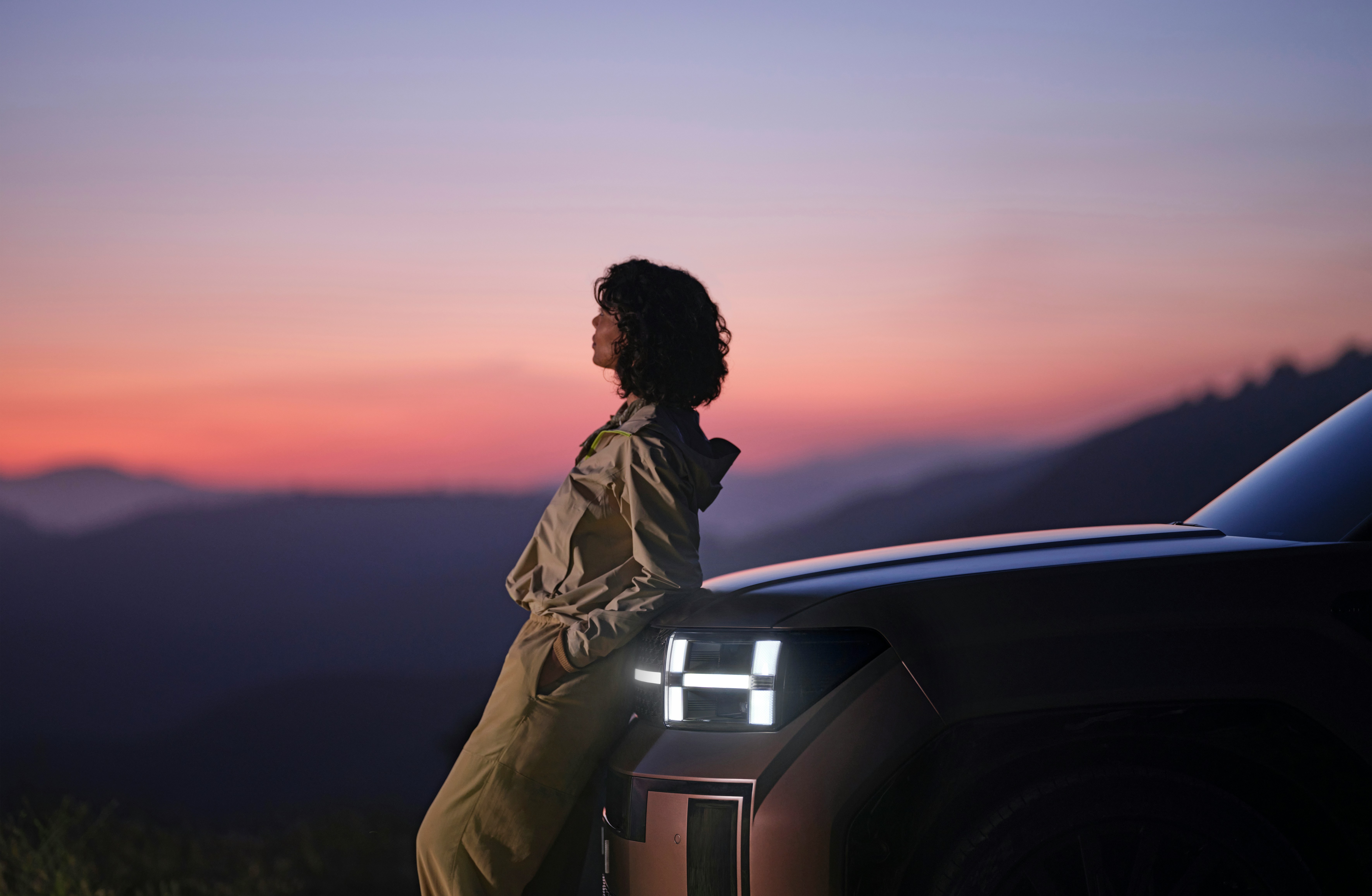 A woman leans back on the hood of an electric vehicle with the dusk sky in the distance.