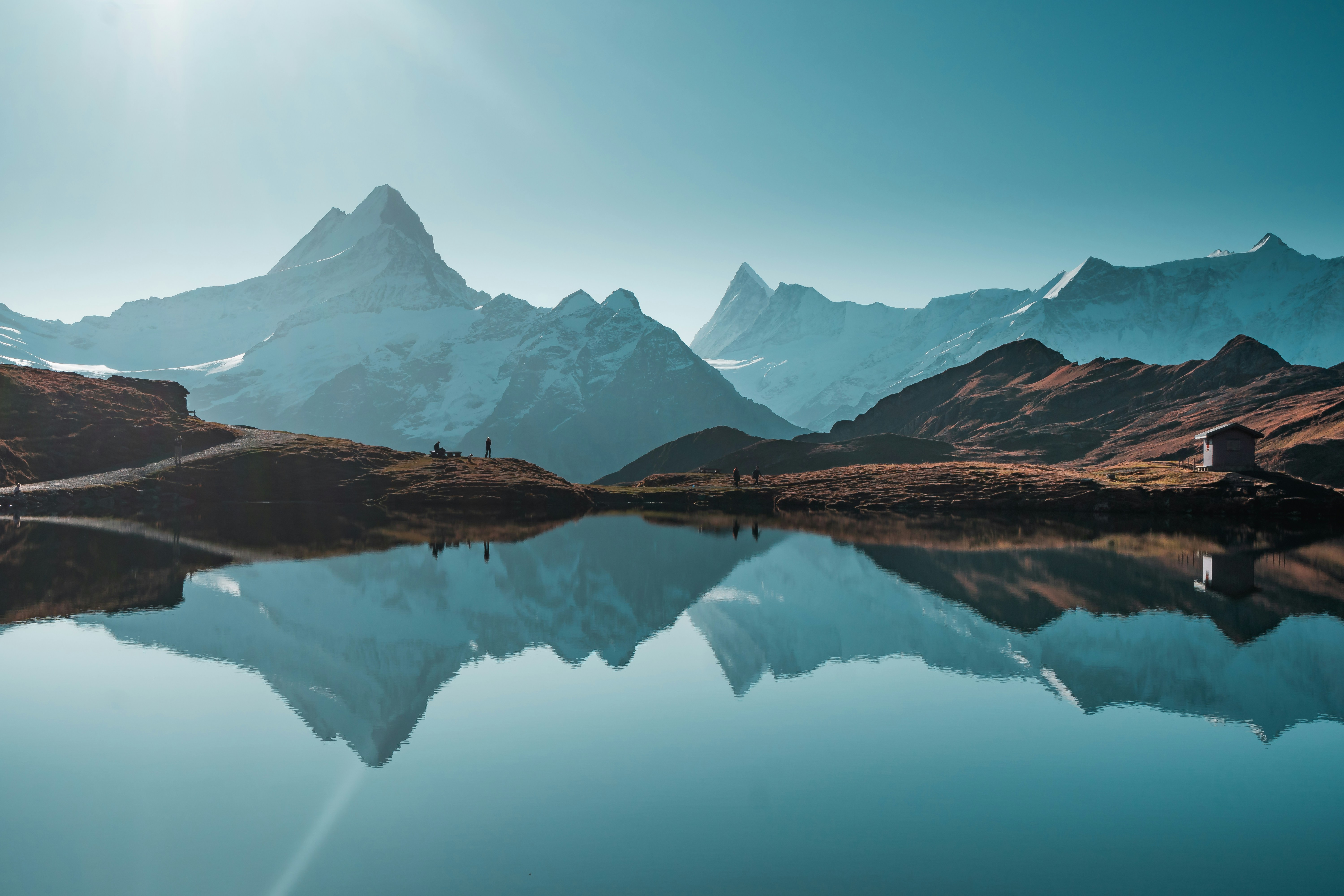 A perfectly flat lake reflects snow-covered mountains in the background with the sun shining.